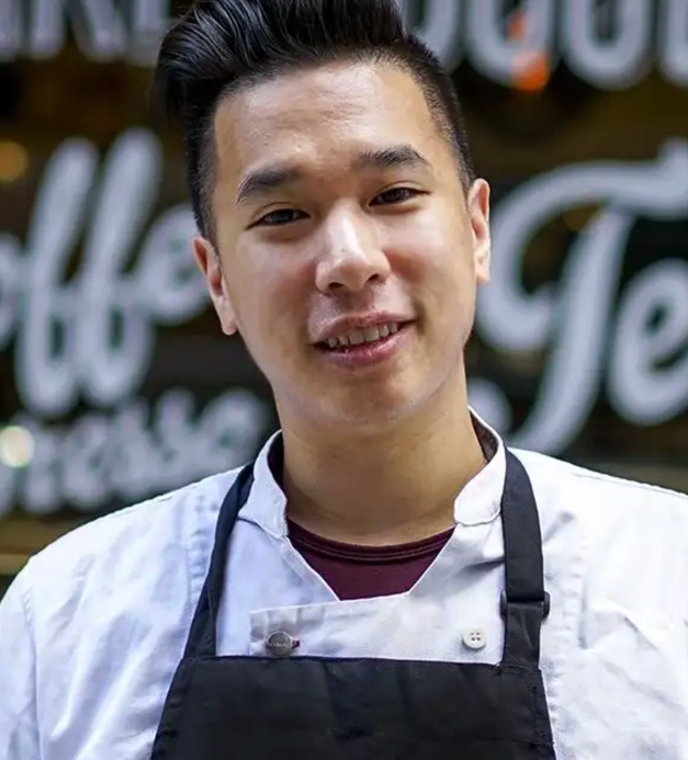 Smiling young Asian man in a white chef's coat and black apron.