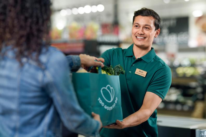 A smiling male store employee hands a "Too Good To Go" bag filled with groceries to a customer.