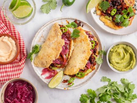 Tacos and a tostada with guacamole, pickled red onions, and a dark filling, surrounded by sauces, limes, and cilantro.