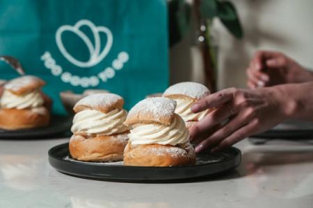 Hands reaching for several cream-filled semlor buns dusted with powdered sugar, with a "Too Good To Go" bag in the background.