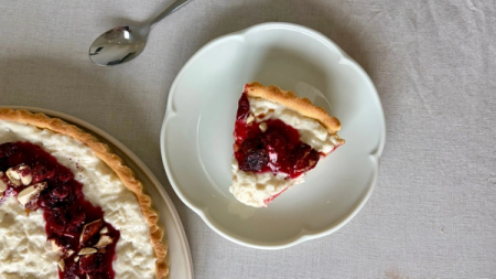 A slice of white tart with red fruit compote and almonds on a white plate, next to a larger matching tart.