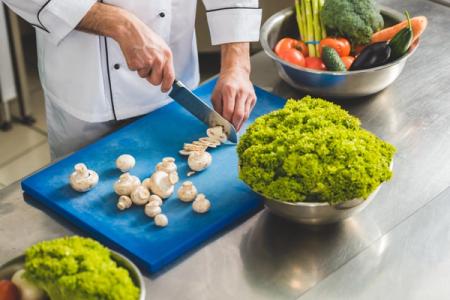 Chef slicing mushrooms on a blue cutting board with fresh vegetables on a metal counter.