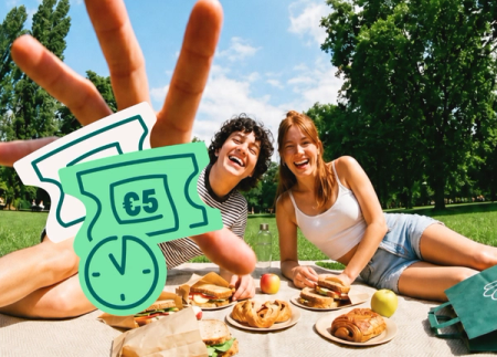 Two laughing friends picnic in a sunny park with food, a hand making a peace sign is above them, and overlaid cartoon €5 discount tickets with a timer icon are visible.
