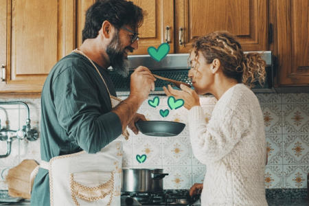 Smiling man in apron feeds woman from a pan with a spoon, with green heart graphics in a kitchen.