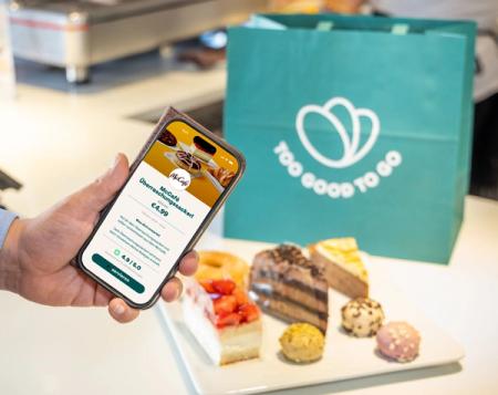 A hand holds a phone displaying the "Too Good To Go" app for a McCafé surprise bag, with a teal Too Good To Go bag and assorted pastries on a plate in the background.