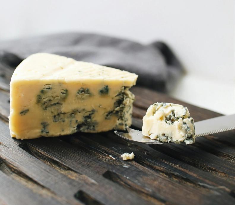 Wedge of blue cheese with a piece on a knife, on a dark wooden board.