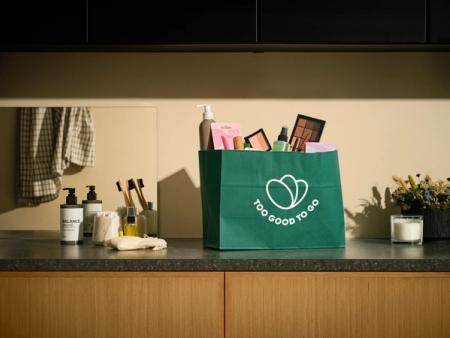 A green "Too Good To Go" bag overflowing with beauty products sits on a bathroom counter.