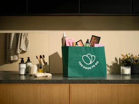A green "Too Good To Go" bag overflowing with beauty products sits on a bathroom counter.