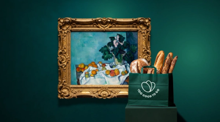 Still life painting of fruit and flowers next to a "Too Good To Go" bag filled with bread.