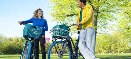 A smiling couple stands with bicycles in a sunny park, both bikes featuring teal bags, with pink hearts floating between them.