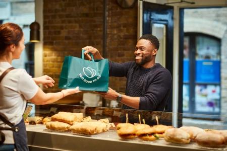 A smiling customer receives a "Too Good To Go" bag from a bakery employee behind a counter of pastries.