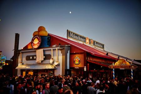 A large, decorated "Münchner Knödelei Biergarten" tent crowded with people at dusk, with a crescent moon in the sky.