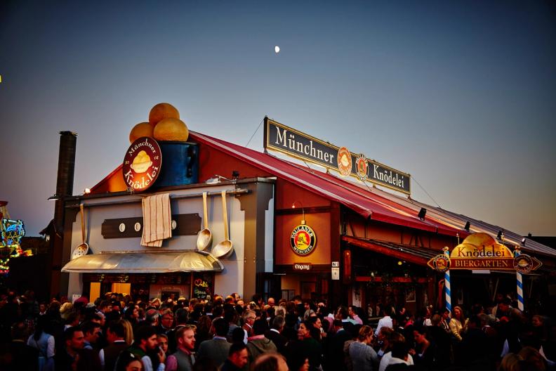 A large, decorated "Münchner Knödelei Biergarten" tent crowded with people at dusk, with a crescent moon in the sky.