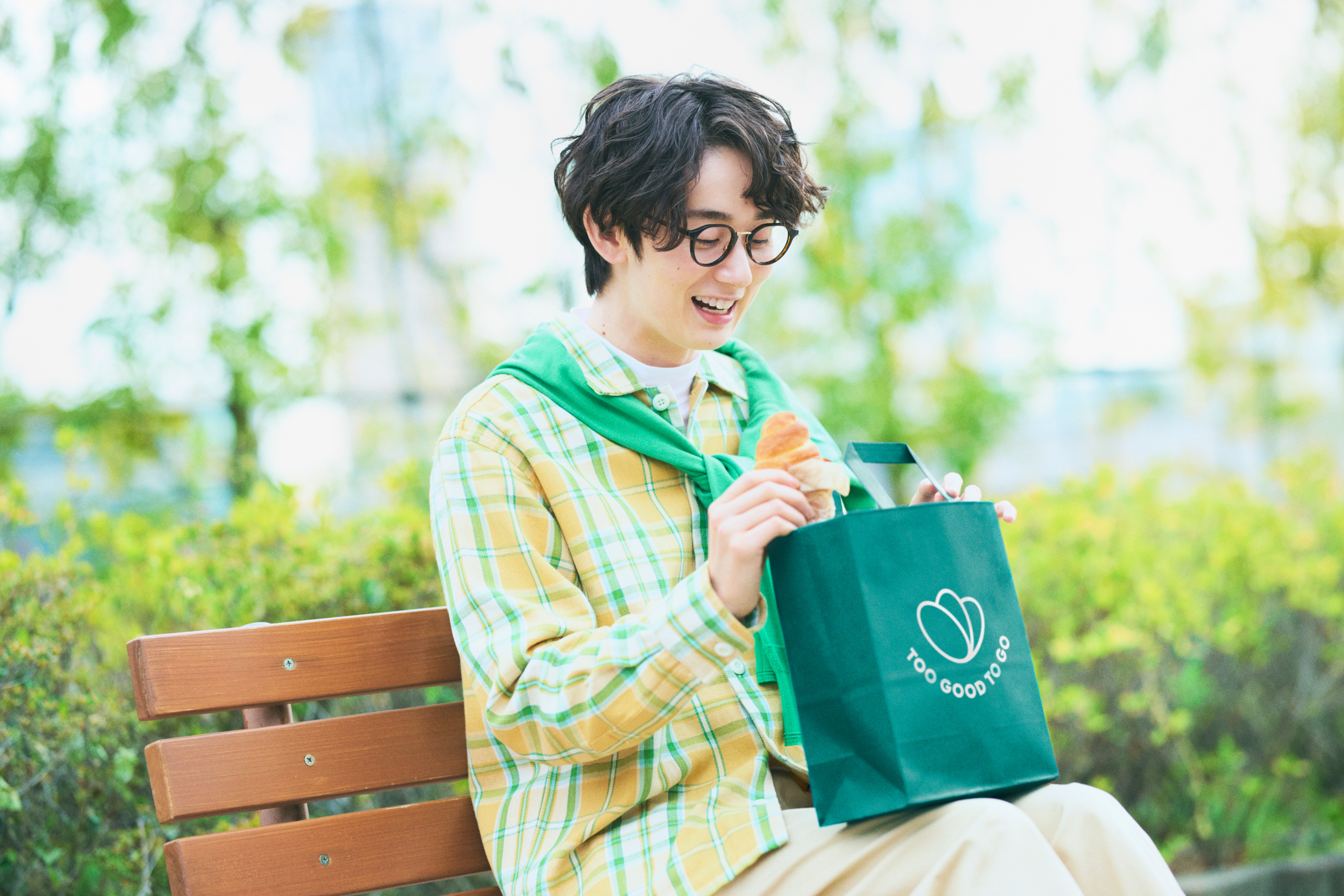 A smiling young man holds a croissant and a "Too Good To Go" bag on a park bench.