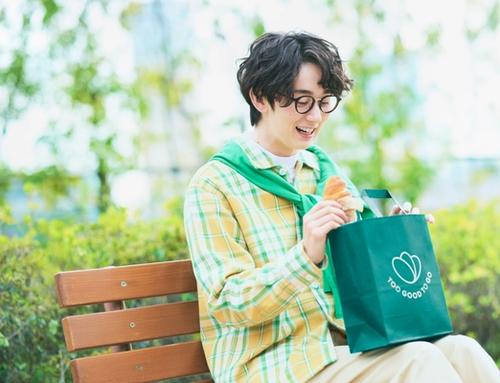 A smiling young man holds a croissant and a "Too Good To Go" bag on a park bench.