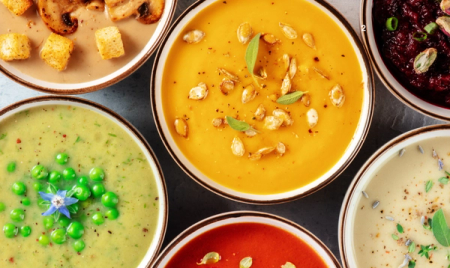 Six bowls of various colorful soups, such as mushroom, pea, and pumpkin.