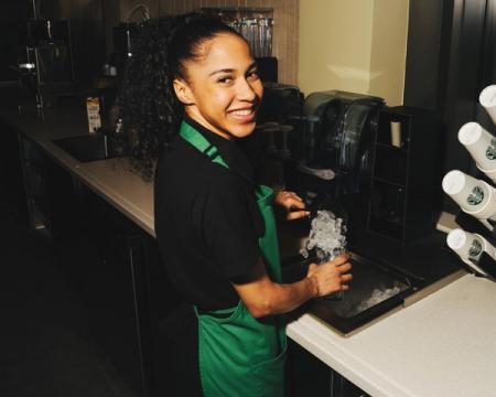A smiling barista in a green apron scoops ice into a cup behind a counter.