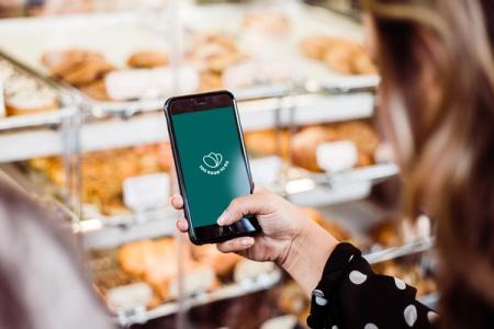 A person holds a smartphone displaying the Too Good To Go app in front of a bakery display case.