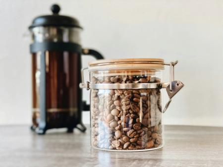 A glass jar filled with coffee beans is sitting on a wooden table next to a french press.