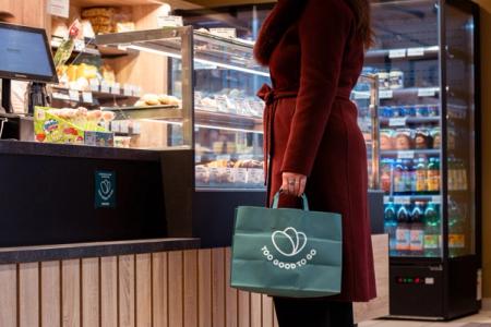 A person in a red coat holds a green "Too Good To Go" bag in a food shop.