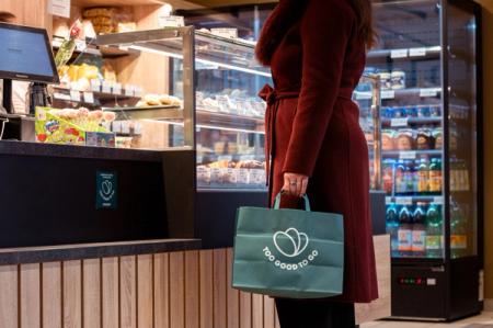 A person in a red coat holds a green "Too Good To Go" bag in a food shop.