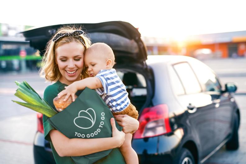 A smiling woman carrying a baby holds a "Too Good To Go" bag full of groceries next to a car with an open trunk in a sunny parking lot.