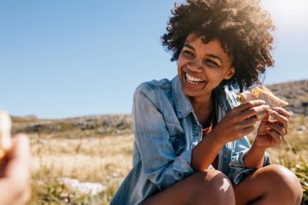 A smiling woman with curly hair eats a sandwich outdoors.
