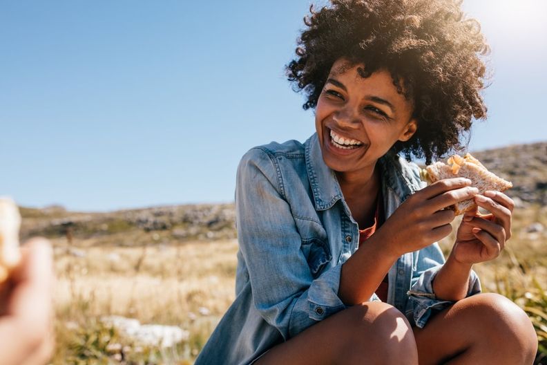A smiling woman with curly hair eats a sandwich outdoors.