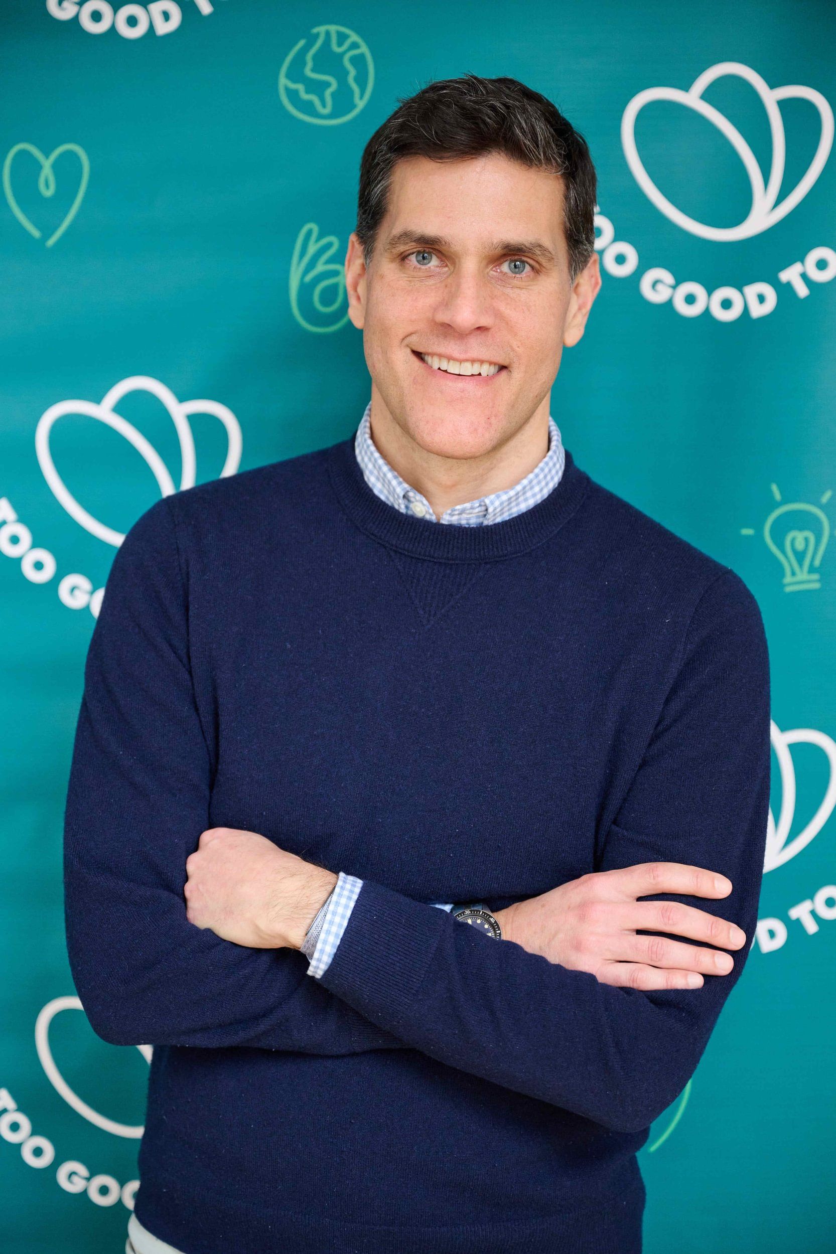 A smiling man in a navy sweater and checkered shirt with arms crossed stands against a teal background with "Too Good To Go" branding.