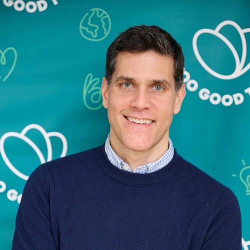 A smiling man in a navy sweater and checkered shirt with arms crossed stands against a teal background with "Too Good To Go" branding.