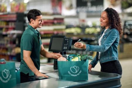 A smiling customer pays for groceries in a "Too Good To Go" bag at a checkout with a smiling cashier.