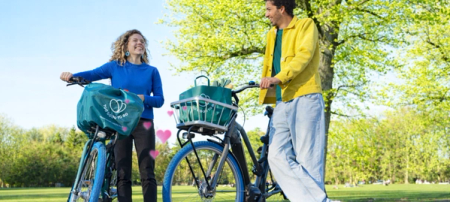 Smiling man and woman with Swapfiets bikes and Too Good To Go bags in a sunny park, with pink hearts between them.