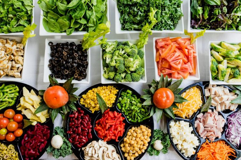 Overhead view of a large salad bar with bowls of fresh ingredients including leafy greens, chopped vegetables, and proteins.