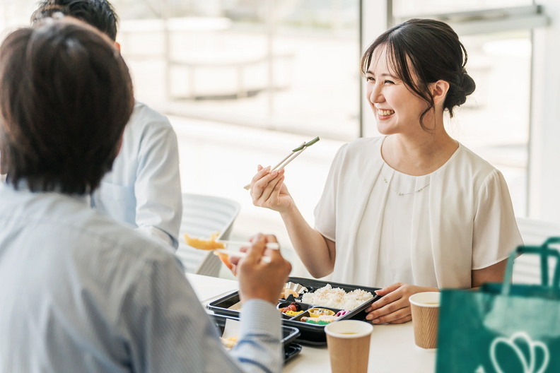 A smiling woman eats a bento box lunch with colleagues.