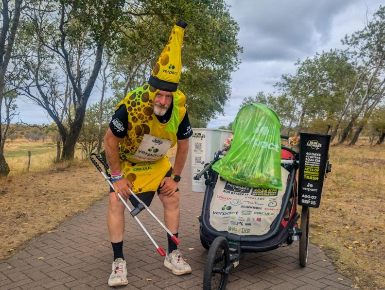A man in a banana costume holds litter pickers next to a cart filled with trash on a paved outdoor path.