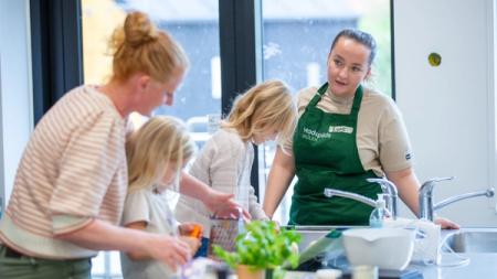A woman and two children in a kitchen, with another woman wearing a green 'Madspilds SKOLEN' (Food Waste School) apron looking on.