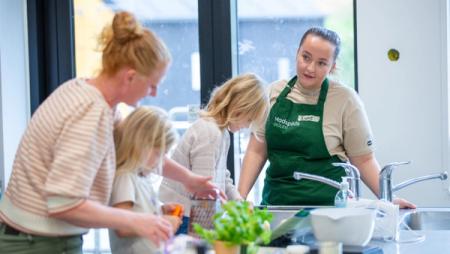 A woman and two children in a kitchen, with another woman wearing a green 'Madspilds SKOLEN' (Food Waste School) apron looking on.