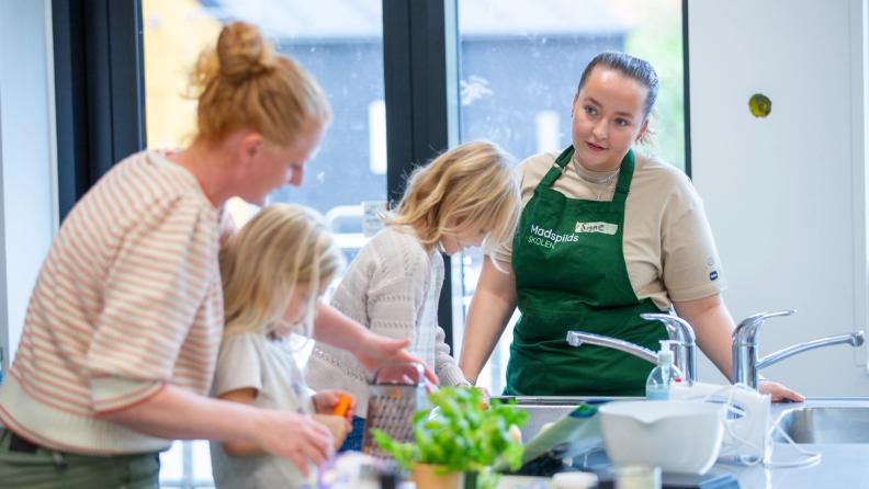A woman and two children in a kitchen, with another woman wearing a green 'Madspilds SKOLEN' (Food Waste School) apron looking on.