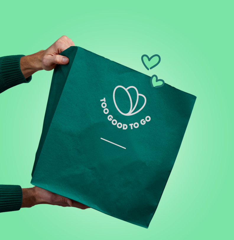 Hands hold a dark green Too Good To Go branded paper bag with its logo and two drawn hearts, on a solid green background.