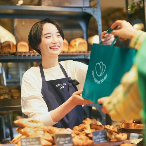 a woman using a tablet next to a bag that says too good to go