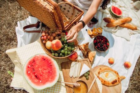 Overhead view of a picnic spread on blankets with a hand reaching for grapes from a basket, surrounded by watermelon, cheese, various fruits, and bread.