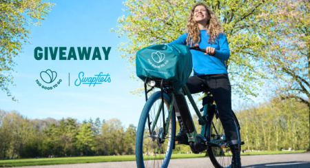 Smiling woman on a bicycle with a 'Too Good To Go' branded basket cover in a sunny park. 'GIVEAWAY' text, Too Good To Go and Swapfiets logos are visible.