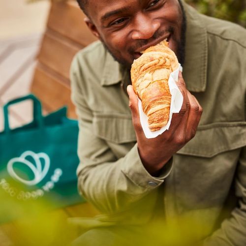 Un homme assis sur un banc dégustant un croissant de son panier Too Good To Go