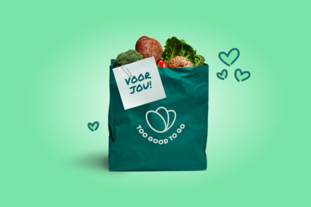 A Too Good To Go Surprise Bag filled with bread and vegetables, a 'VOOR JOU!' note clipped on, against a light green background with hearts.