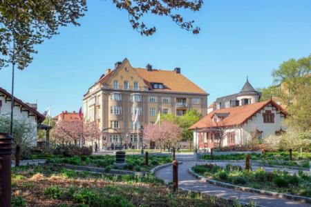 A spring park with cherry blossoms and winding paths, bordered by historic buildings with red roofs under a clear blue sky.