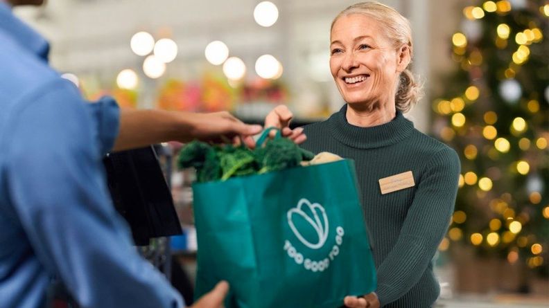 A smiling woman hands a green "Too Good To Go" bag filled with vegetables to a customer, with a decorated Christmas tree in the background.