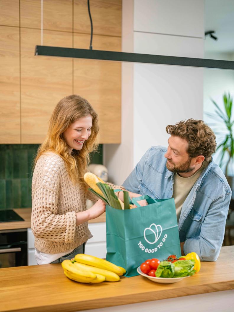 a man and woman in a kitchen with a bag that says too good to go