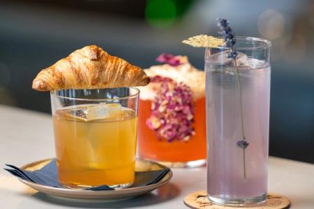 Three cocktails on a bar: a golden drink garnished with a croissant, a red drink with rose petals, and a purple drink with lavender.