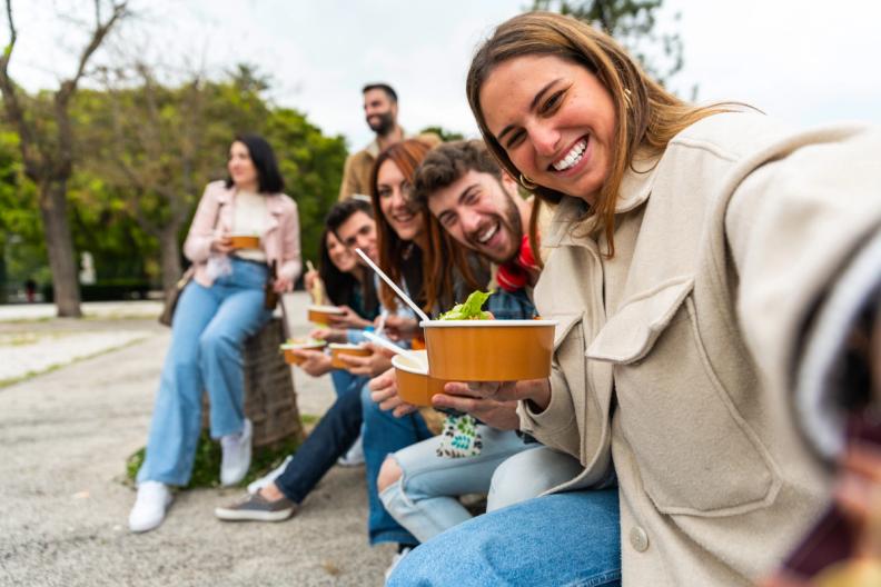 a group of young people are sitting on a bench eating food and taking a selfie .