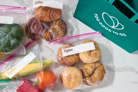Broccoli, corn, bell peppers, pastries, and bagels packaged in Ziploc bags, next to a green "Too Good To Go" bag.